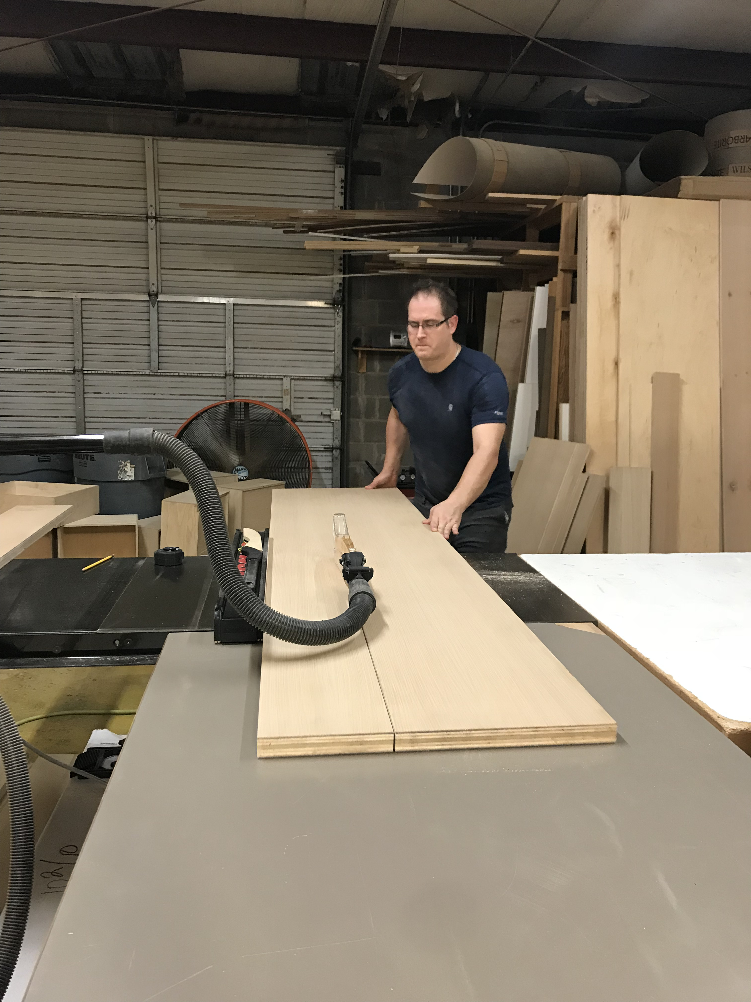 Joel Hodge in a workshop guiding a light wood panel across a worktable with sheet goods and equipment nearby.