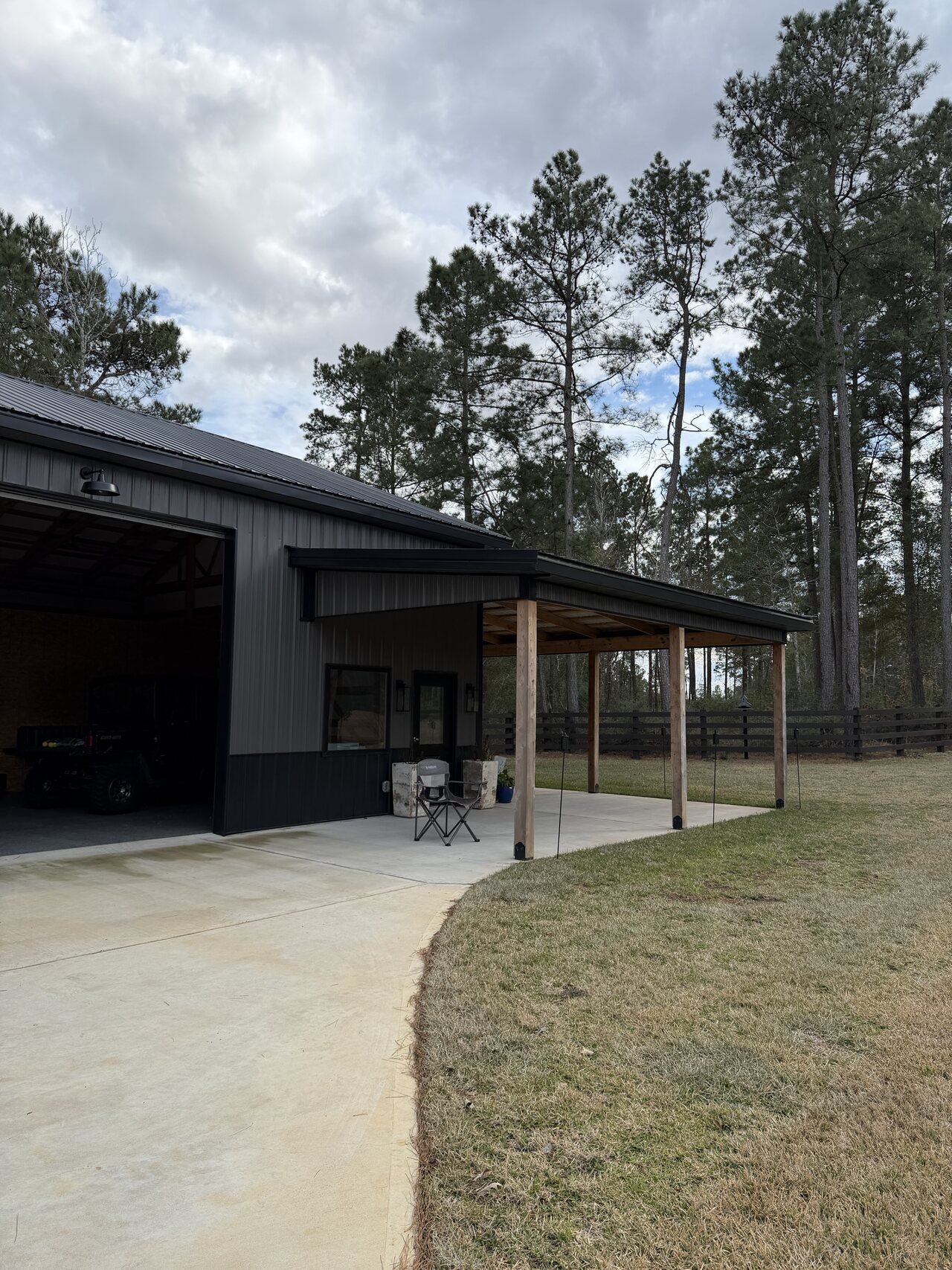 Exterior approach view of a dark metal barn with covered concrete apron and wooded setting.