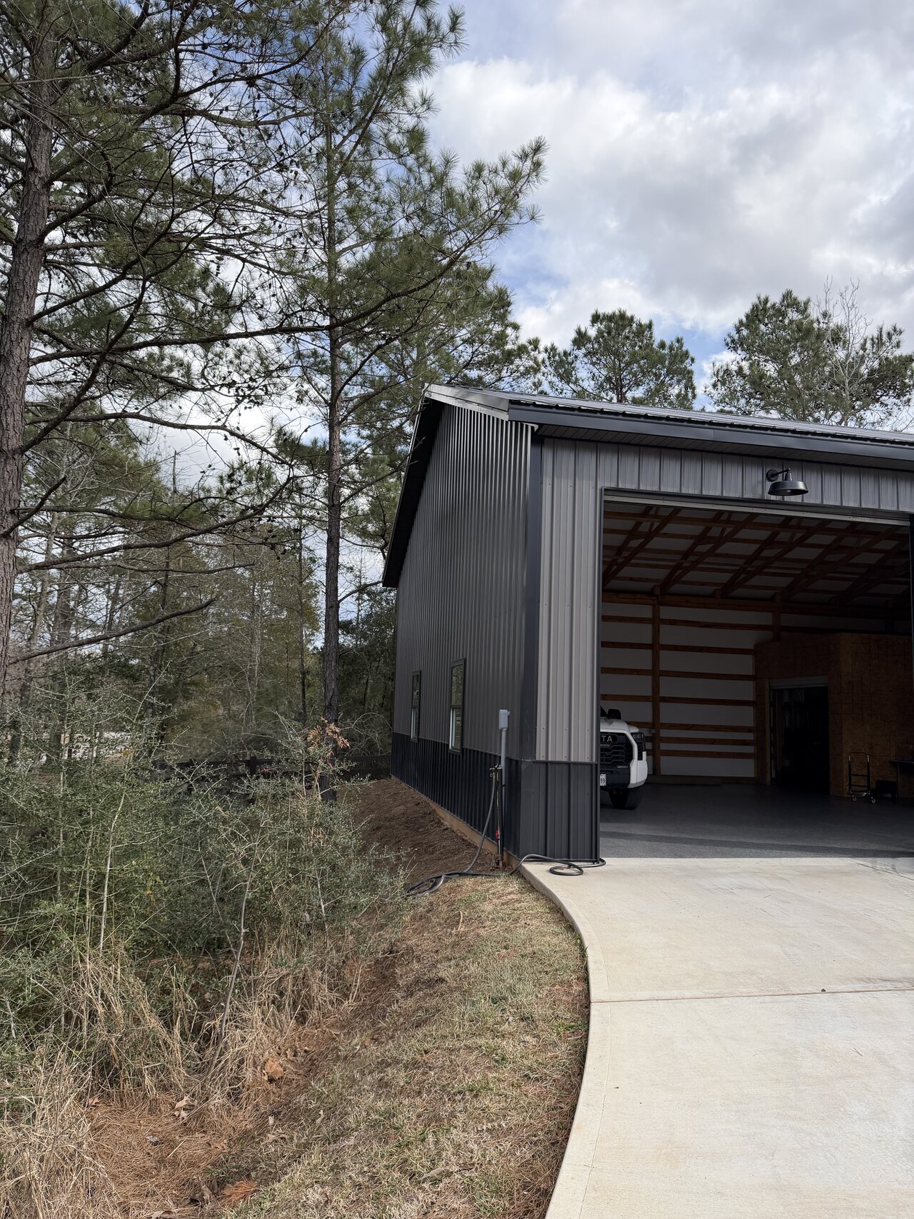 Side exterior view of the dark metal barn with roof overhang, concrete access, and trees.