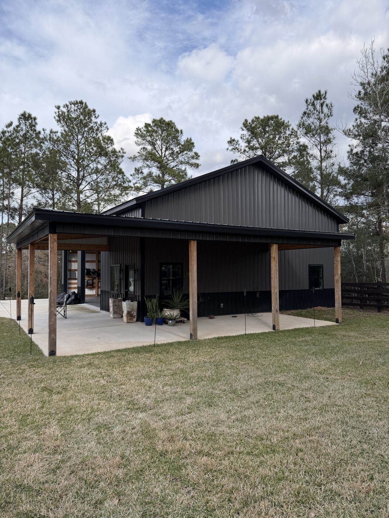 Completed dark metal barn exterior with covered concrete apron, wood posts, black trim, and tree line.