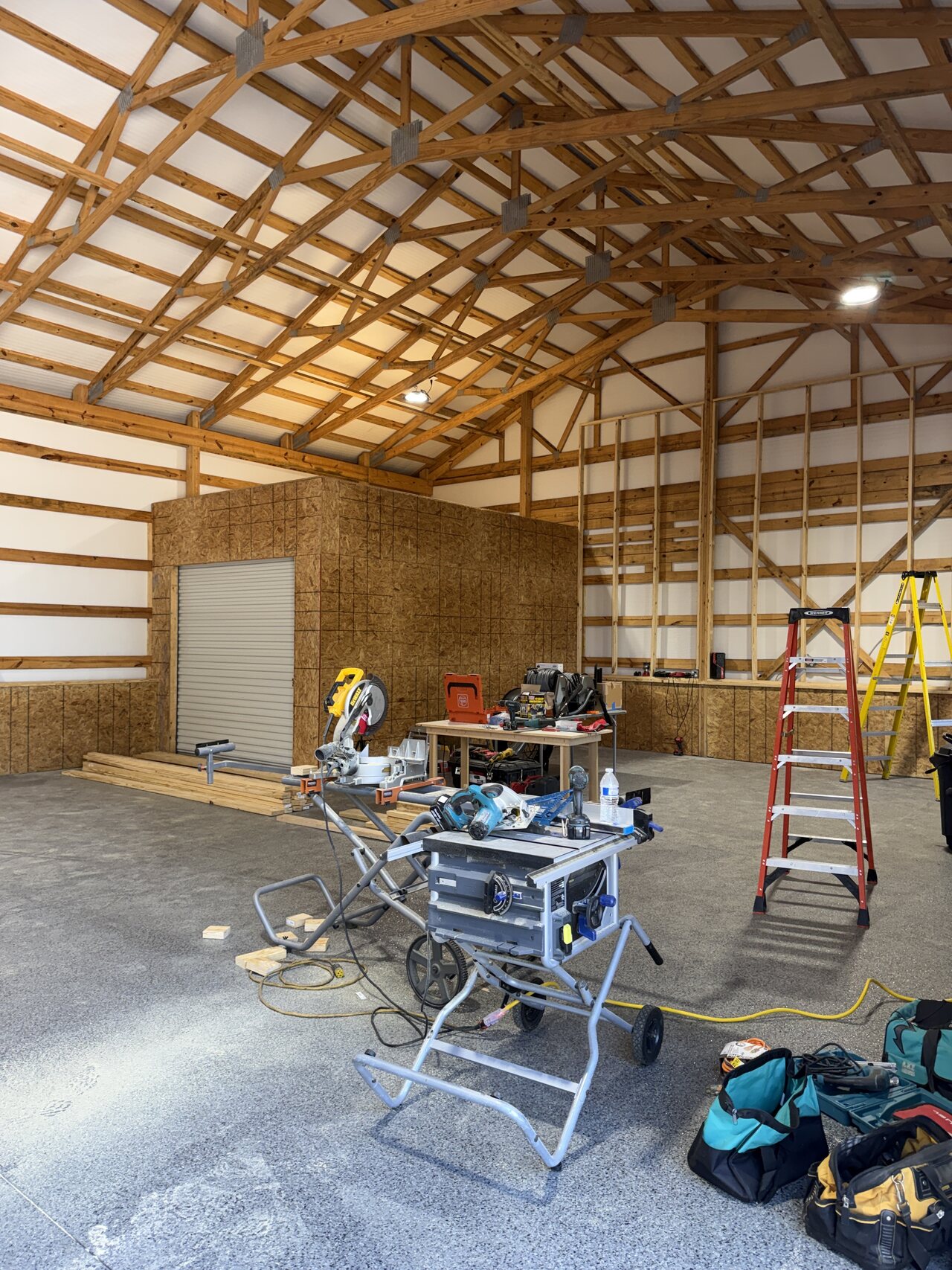 Interior barn work area with saws, ladders, OSB enclosure, exposed trusses, and staged tools.