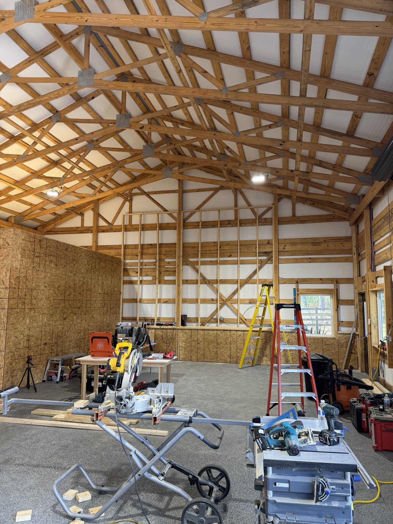Wide in-progress barn interior with roof trusses, OSB enclosure, framed wall section, and ladders.
