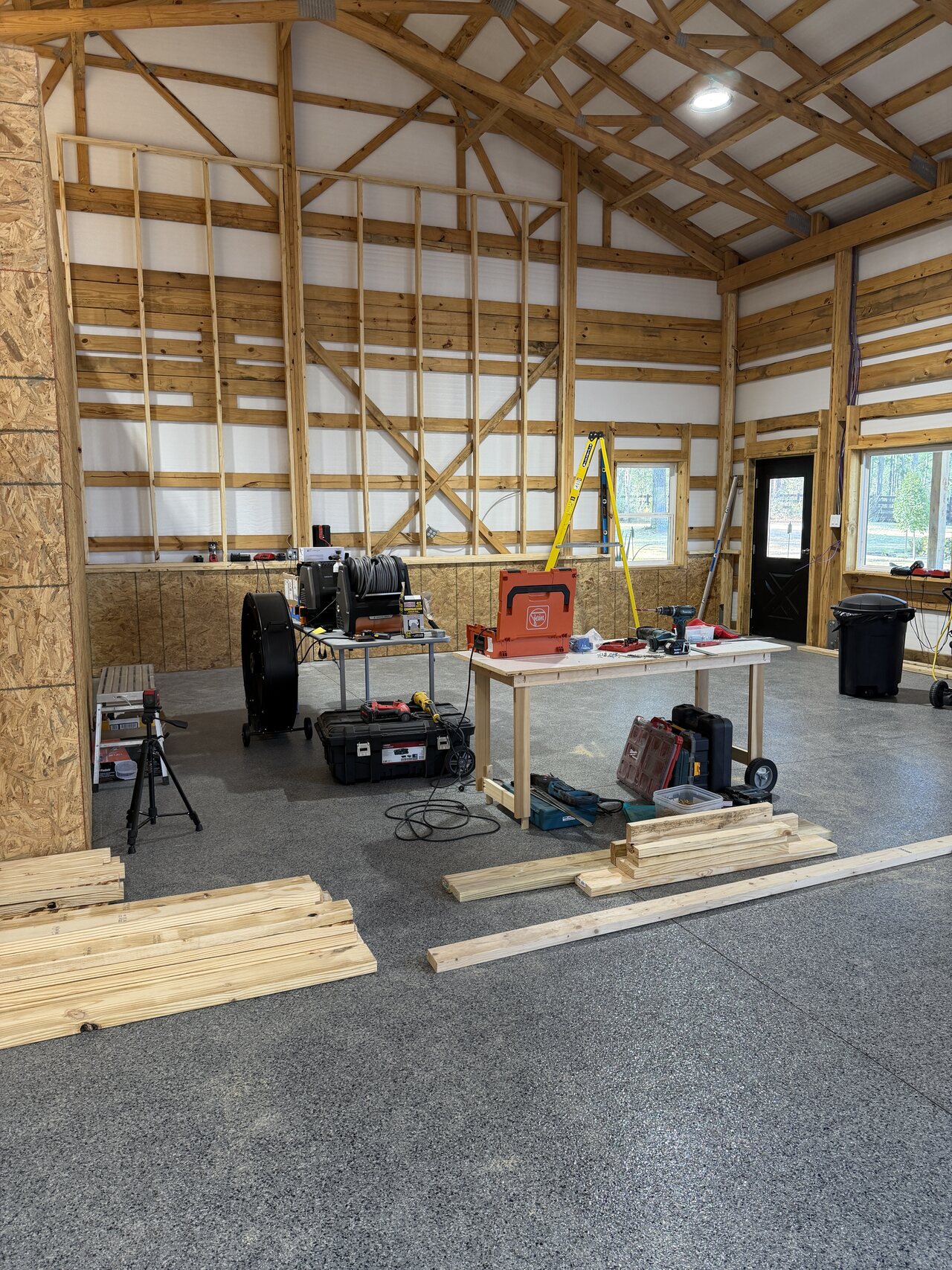 In-progress barn interior with staged tools, open floor, high trusses, and wall woodwork underway.