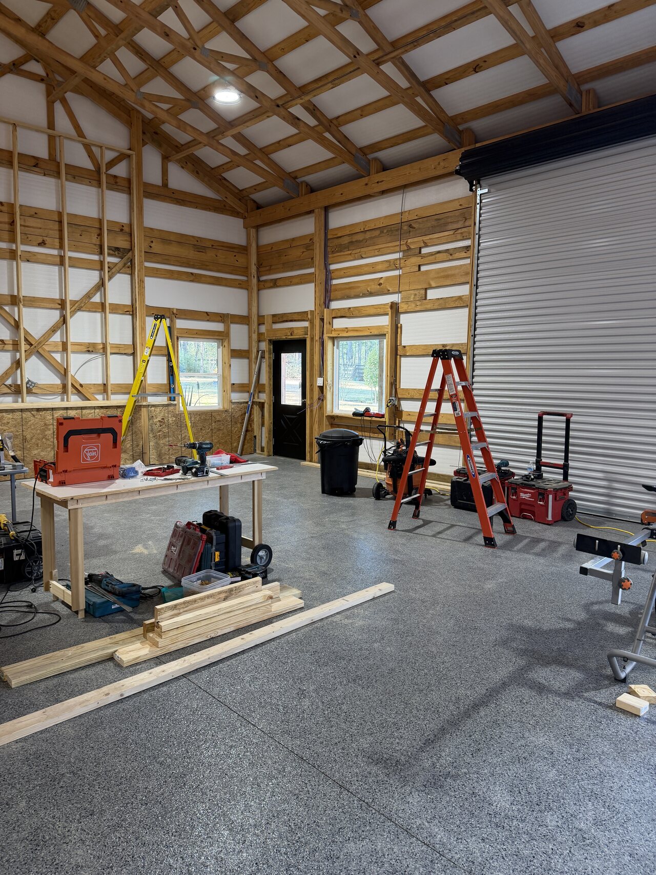 In-progress barn wall work with ladder, tools, roll-up enclosure, and exposed wood trusses.