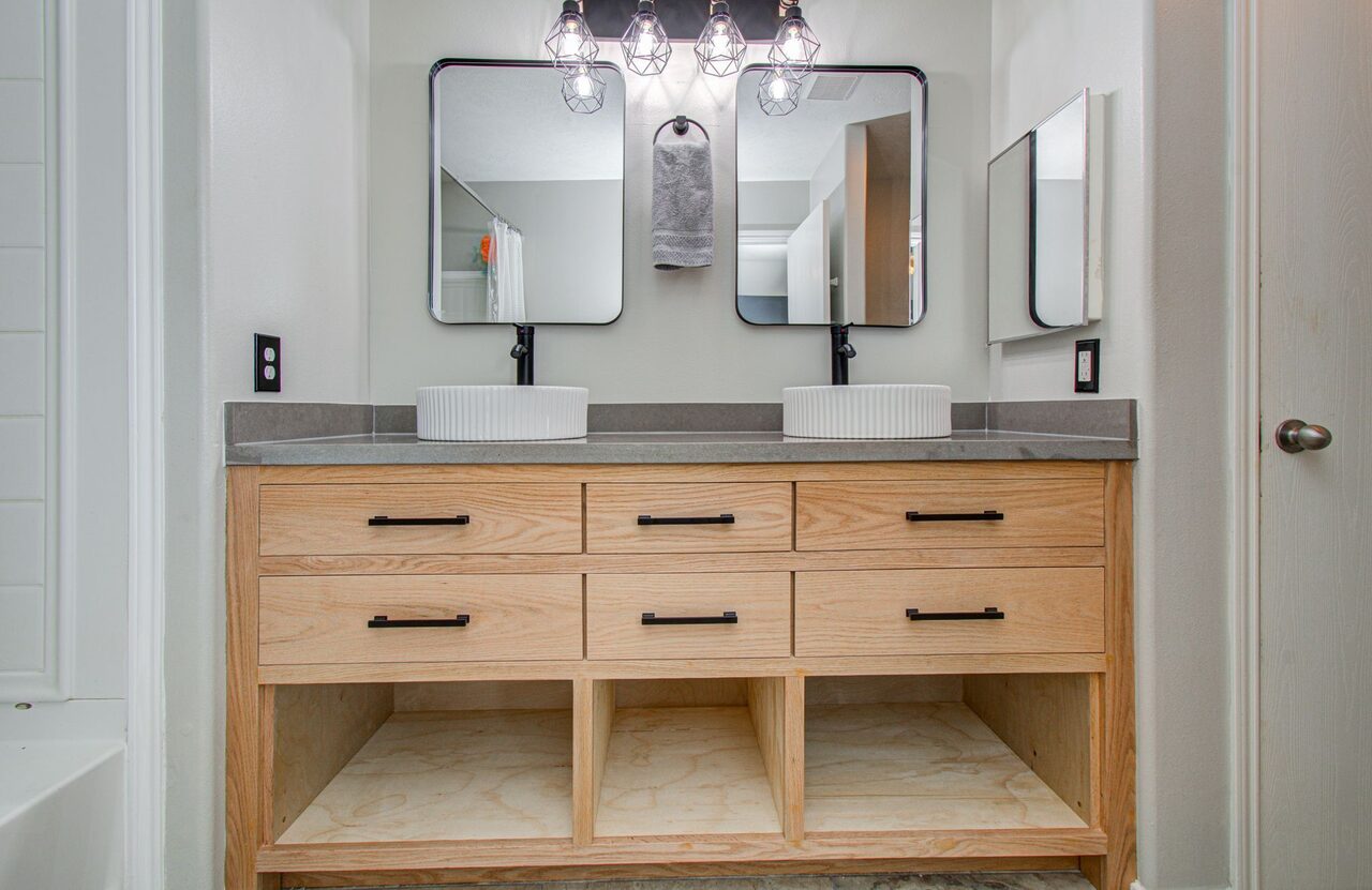 Close view of the installed light wood vanity cabinets, drawer stack, mirror spacing, and lower shelf openings for a Joel Hodge project.