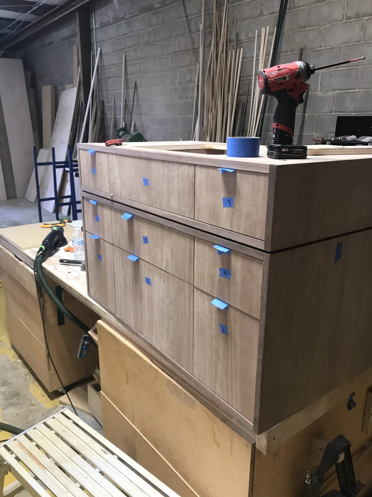 Light wood drawer faces and taped pulls reviewed on the bench before final hardware installation for a Joel Hodge project.