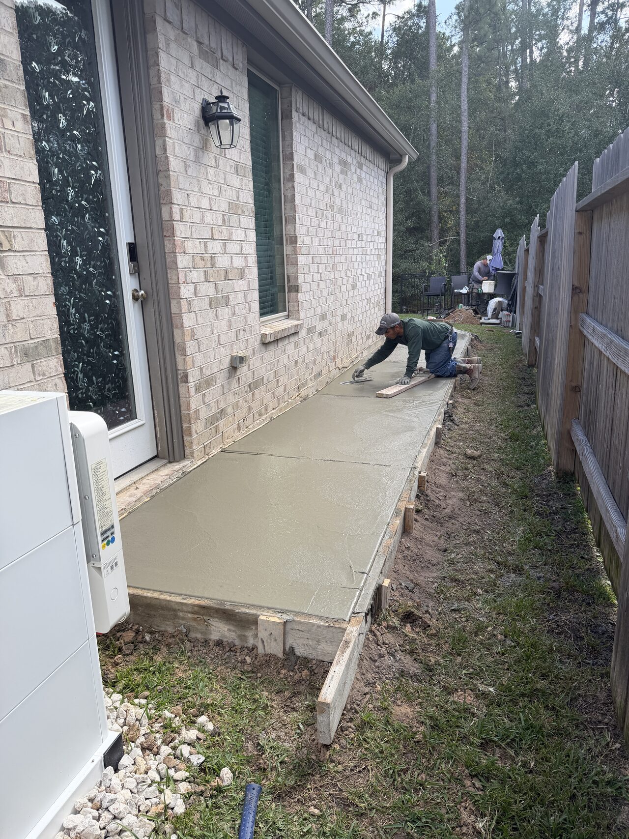 Fresh side-yard concrete path along the brick wall after placement and edge cleanup for a Joel Hodge project.